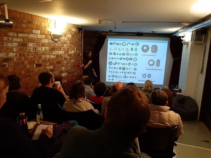 Photo taken from the back of a room showing a seated audience looking towards a spaker and a projected powerpoint slide showing different archaeological beads