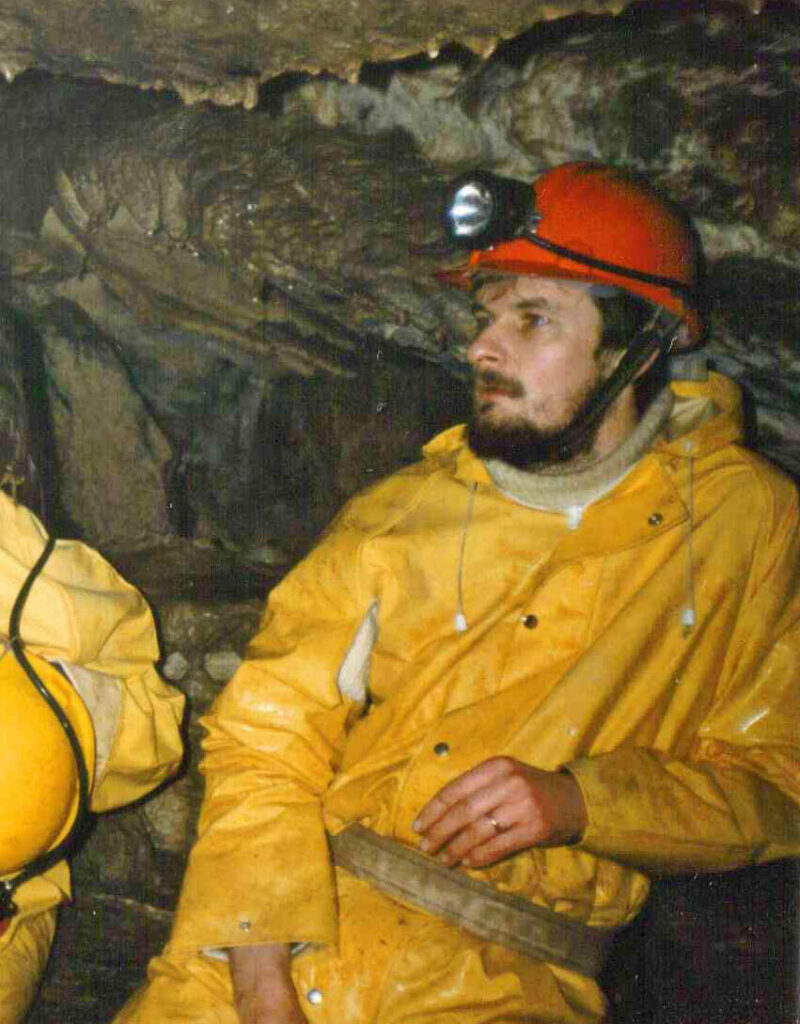 A young geologist wearing a bright yellow waterproof suit and an orange hardhat with a headlamp leans against the wall of a dim cave.